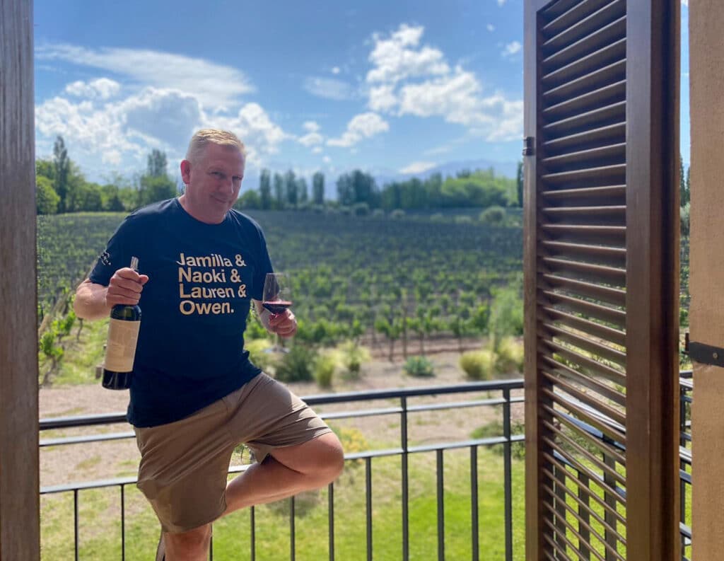 Yuri Koster holding a wine bottle and a glass of red wine while standing on a balcony with Casarena vineyards in the background.