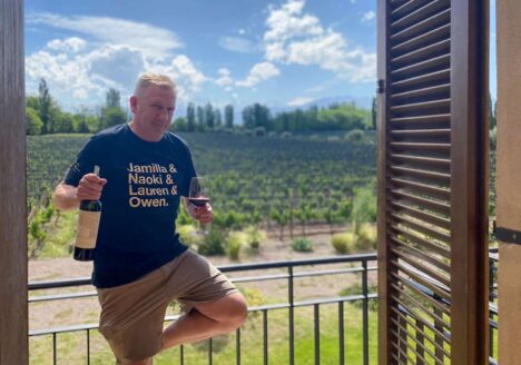 Yuri Koster holding a wine bottle and a glass of red wine while standing on a balcony with Casarena vineyards in the background.